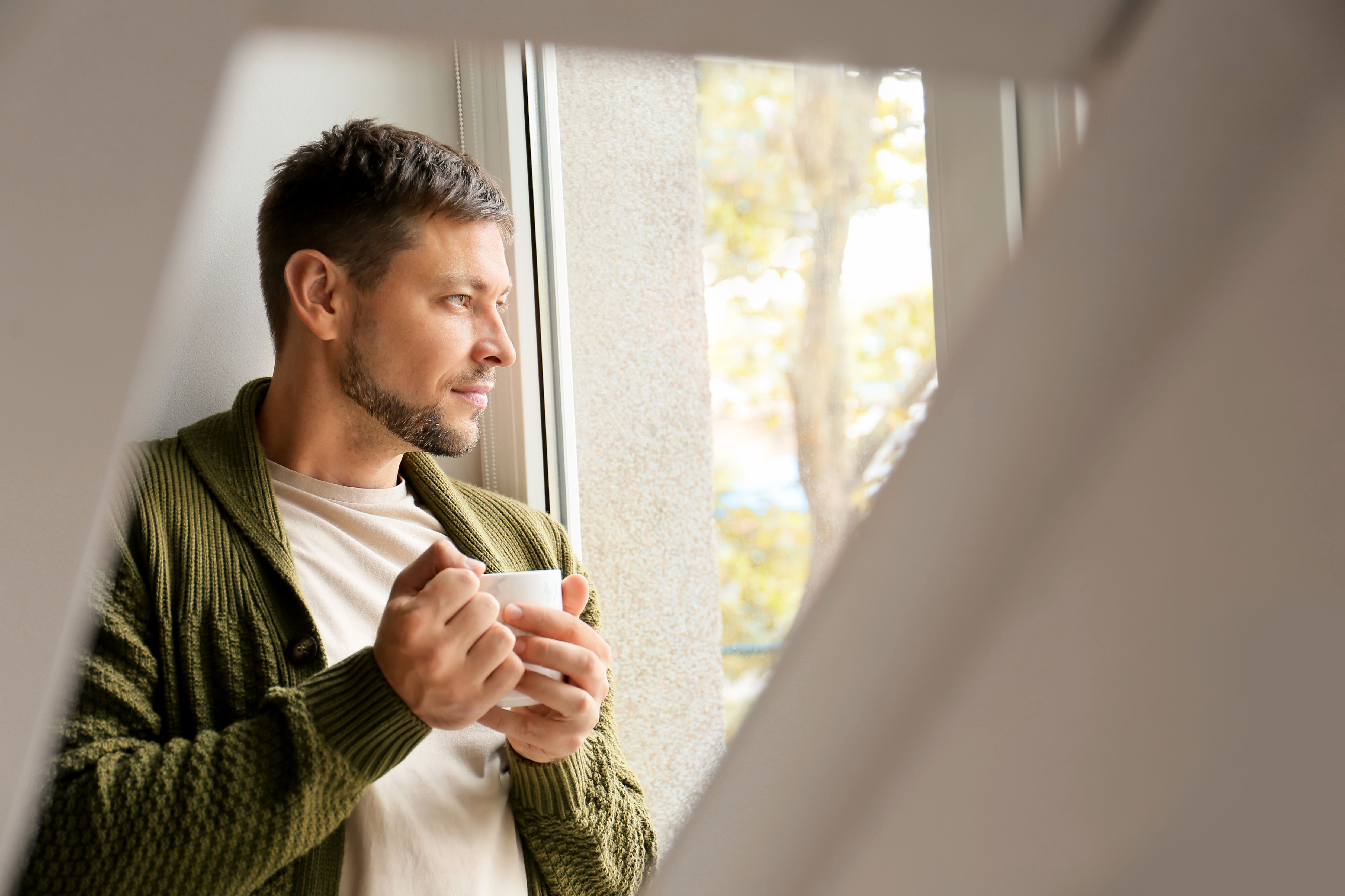 Mann steht mit einer Tasse am Fenster und schaut nachdenklich hinaus